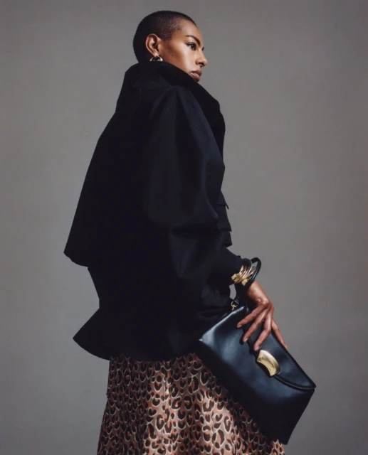 Adult female in a black jacket poses with a black clutch purse and a leopard print skirt against a plain studio backdrop.
