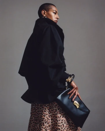 Adult female in a black jacket poses with a black clutch purse and a leopard print skirt against a plain studio backdrop.