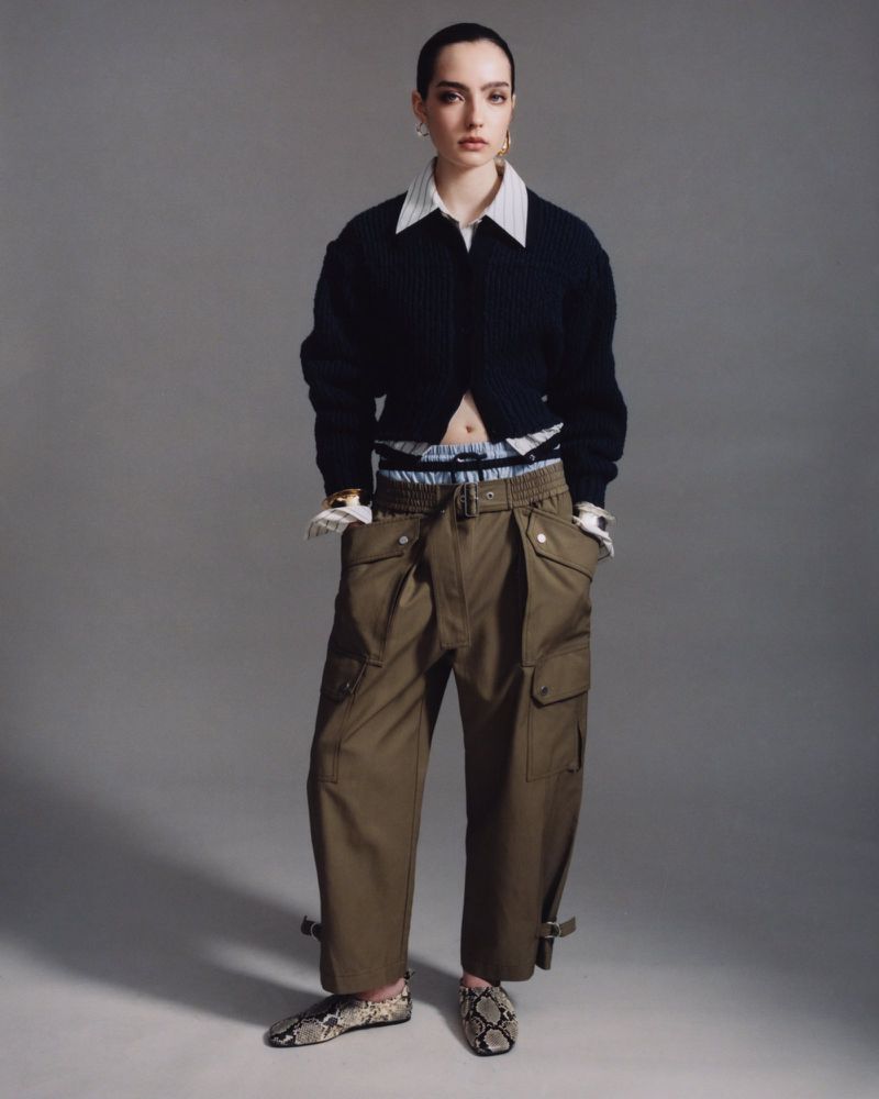 Fashionable adult female in a blue cardigan, olive green pants, and snakeskin shoes poses in a studio setting with a gray background.