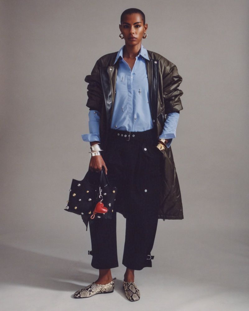 Full-length portrait of a woman wearing a blue button-down shirt, black pants, a long olive green coat, and patterned shoes with a black tote bag.