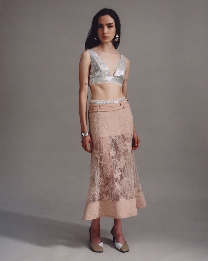 Full-length shot of a woman posing in a silver V-neck top and a peach lace skirt in a studio with a light grey backdrop.