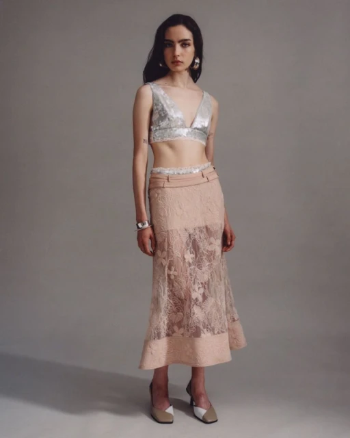 Full-length shot of a woman posing in a silver V-neck top and a peach lace skirt in a studio with a light grey backdrop.