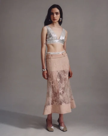 Full-length shot of a woman posing in a silver V-neck top and a peach lace skirt in a studio with a light grey backdrop.