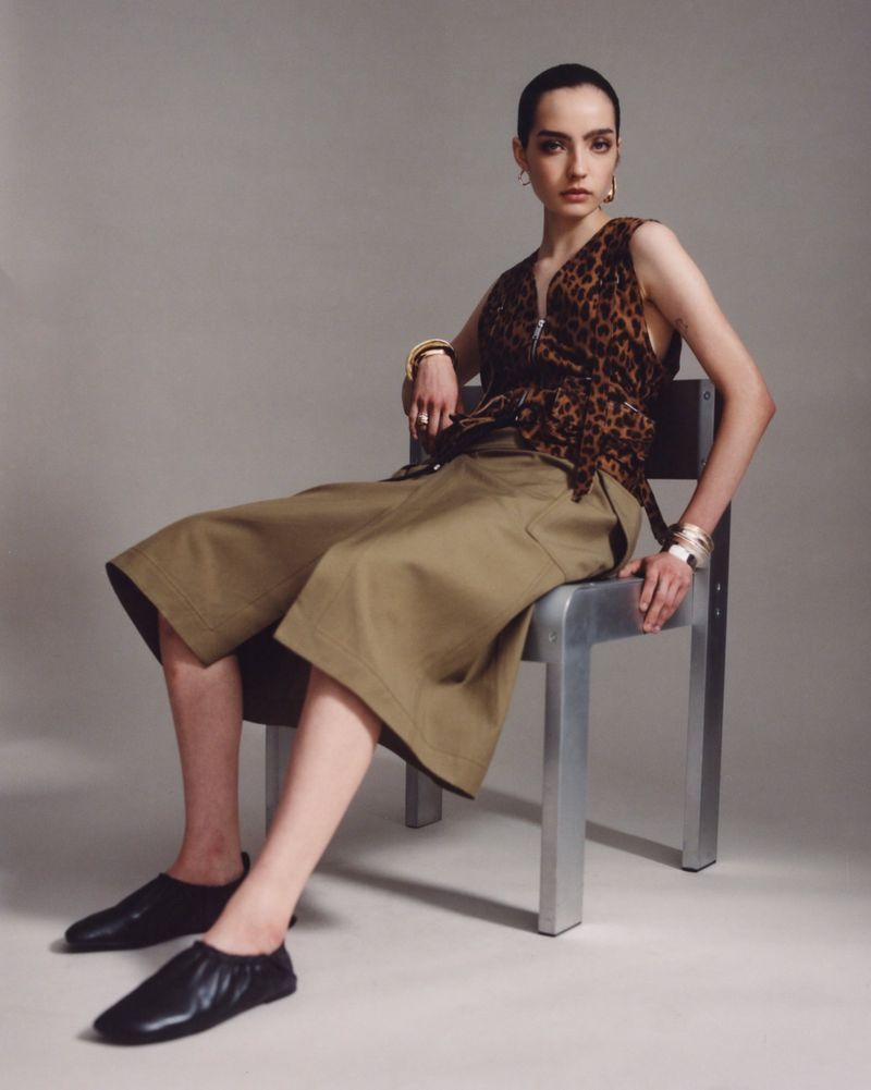 A brunette woman in a leopard-print top and olive green culottes is seated on a chrome chair in a studio setting. Fashion shot.