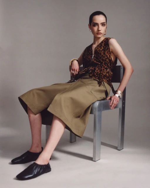 A brunette woman in a leopard-print top and olive green culottes is seated on a chrome chair in a studio setting. Fashion shot.