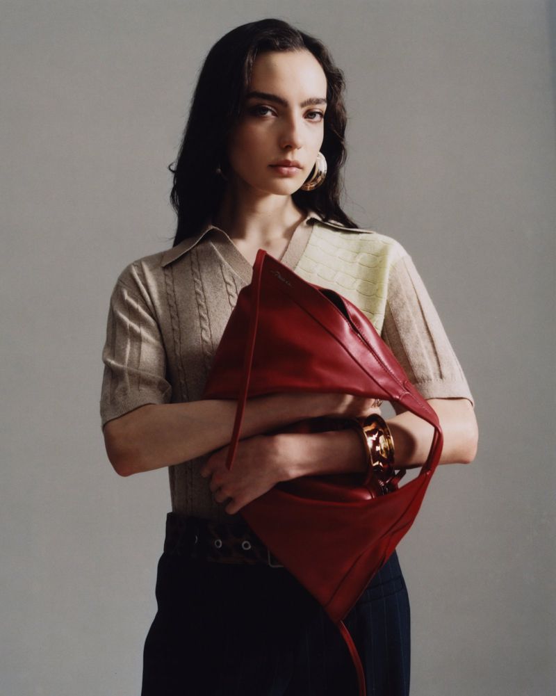 Model posing with a large red leather shoulder bag and beige knit polo shirt against a plain grey studio backdrop.