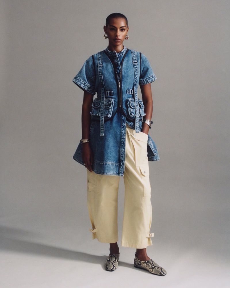 Fashionable woman wearing denim dress with pockets, khaki trousers, and snakeskin shoes in a full-length studio shot on grey.