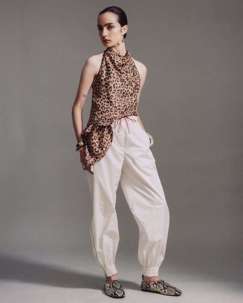 A young woman in a leopard-print halter top and white pants strikes a full-length pose in a studio, highlighted by neutral gray background.