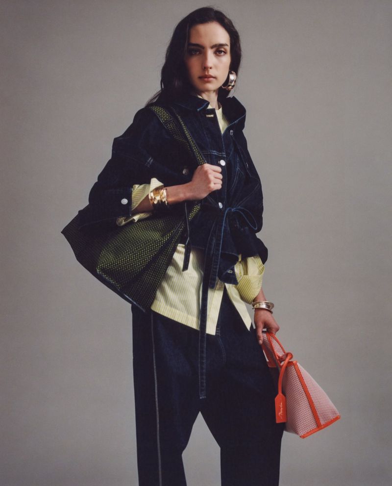 Brunette woman in denim jacket, oversized trousers and light shirt holding two tote bags in a studio setting.