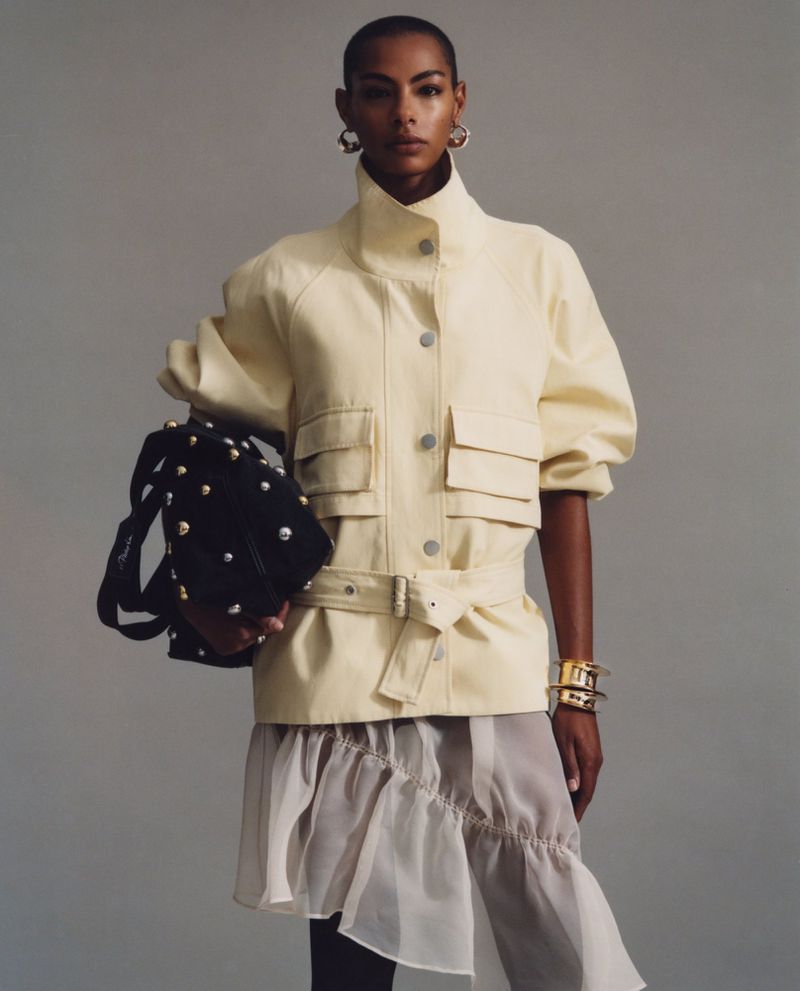 Fashion editorial shot of a Black female model wearing a belted jacket, layered skirt, and accessories, holding a studded handbag.