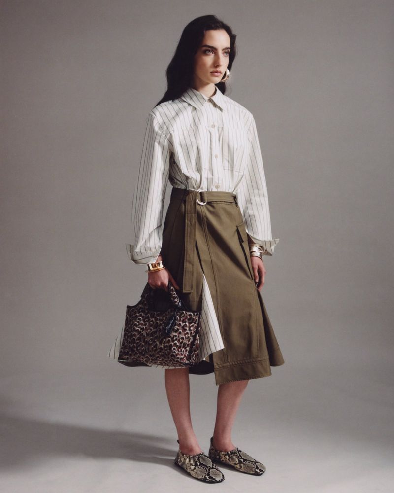 Fashion photo of a stylish woman in a striped shirt and khaki skirt, accessorized with a leopard bag and snake print shoes against a grey backdrop.