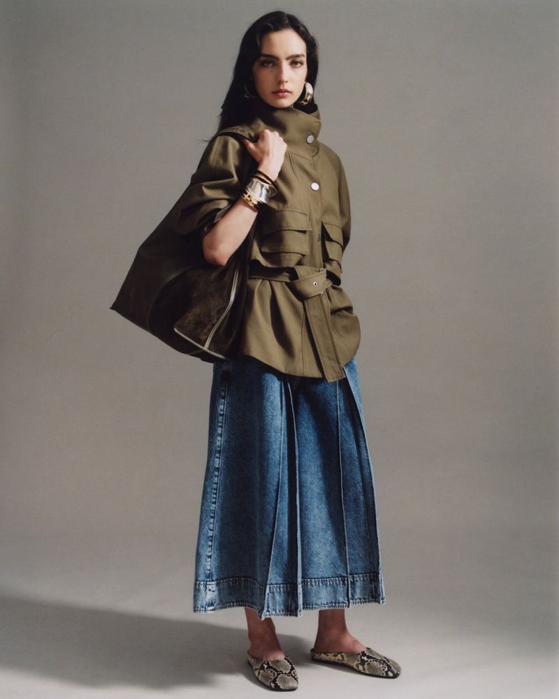 Fashion shot: Brunette woman in an olive jacket and wide leg jeans holding a bag. She is wearing statement earrings and snake print flats.