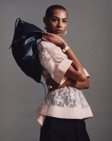 Model with buzzcut wearing a sheer blush lace top and black trousers holding a slouchy black bag against a grey studio backdrop.