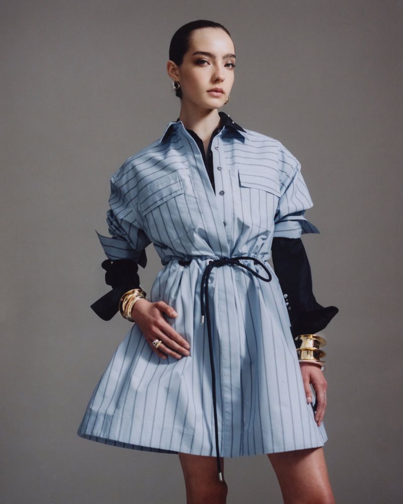Young woman in a light blue striped dress with a black belt and stacked gold bracelets posing in a studio with a grey background.