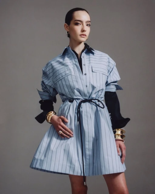 Young woman in a light blue striped dress with a black belt and stacked gold bracelets posing in a studio with a grey background.