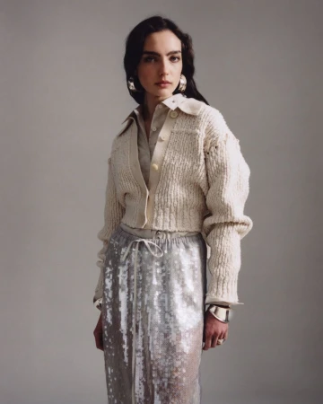 Portrait of a woman with dark hair, wearing a cream-colored knit cardigan, silver sequin skirt, large hoop earrings, and a silver bracelet.