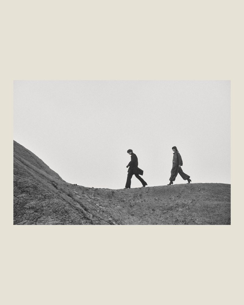 Two fashion models in dark winter outerwear walking along a rocky ridgeline against an overcast sky in black and white.