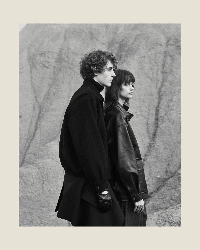 A young man with curly hair and a woman with bangs standing side-by-side wearing black jackets against a textured rock formation.