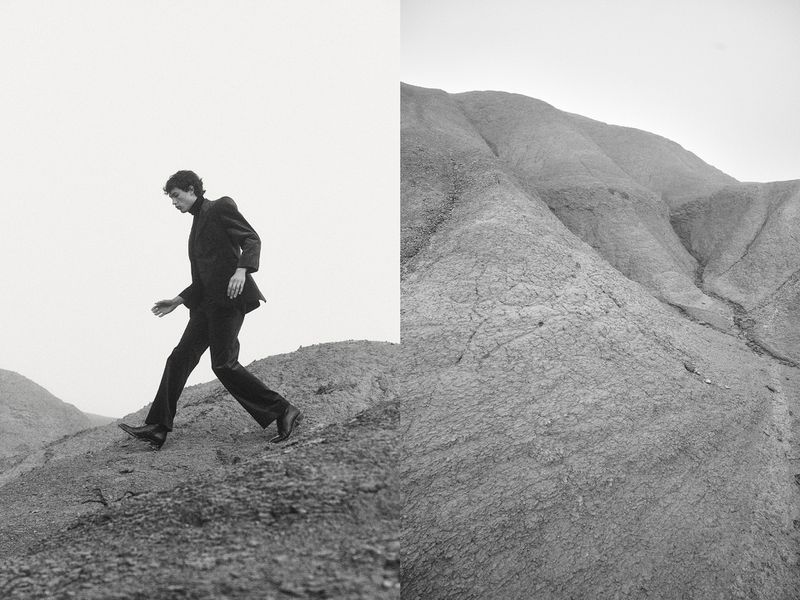 Model wearing a dark tailored suit and leather boots walking across a barren, rocky hillside in a black and white fashion composition.