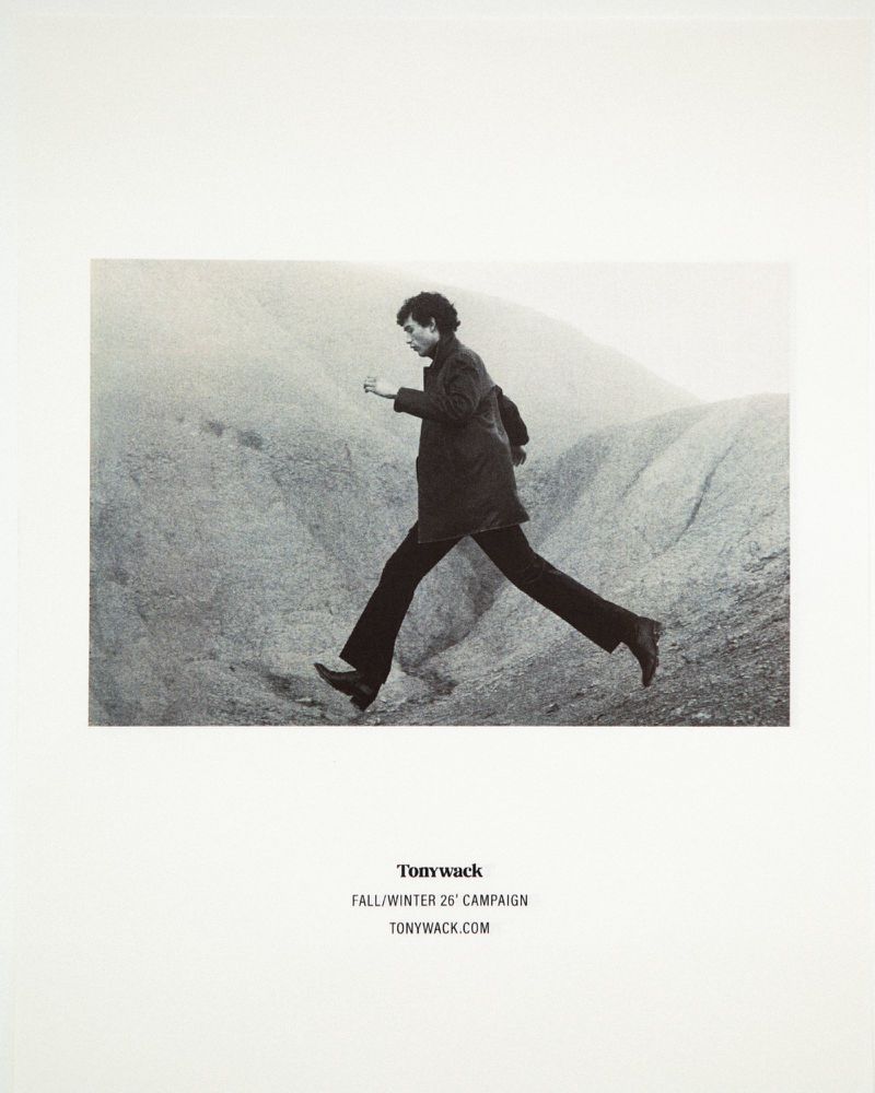 Monochrome fashion campaign poster showing a model leaping mid-air across a rugged hillside in a dark jacket and trousers.