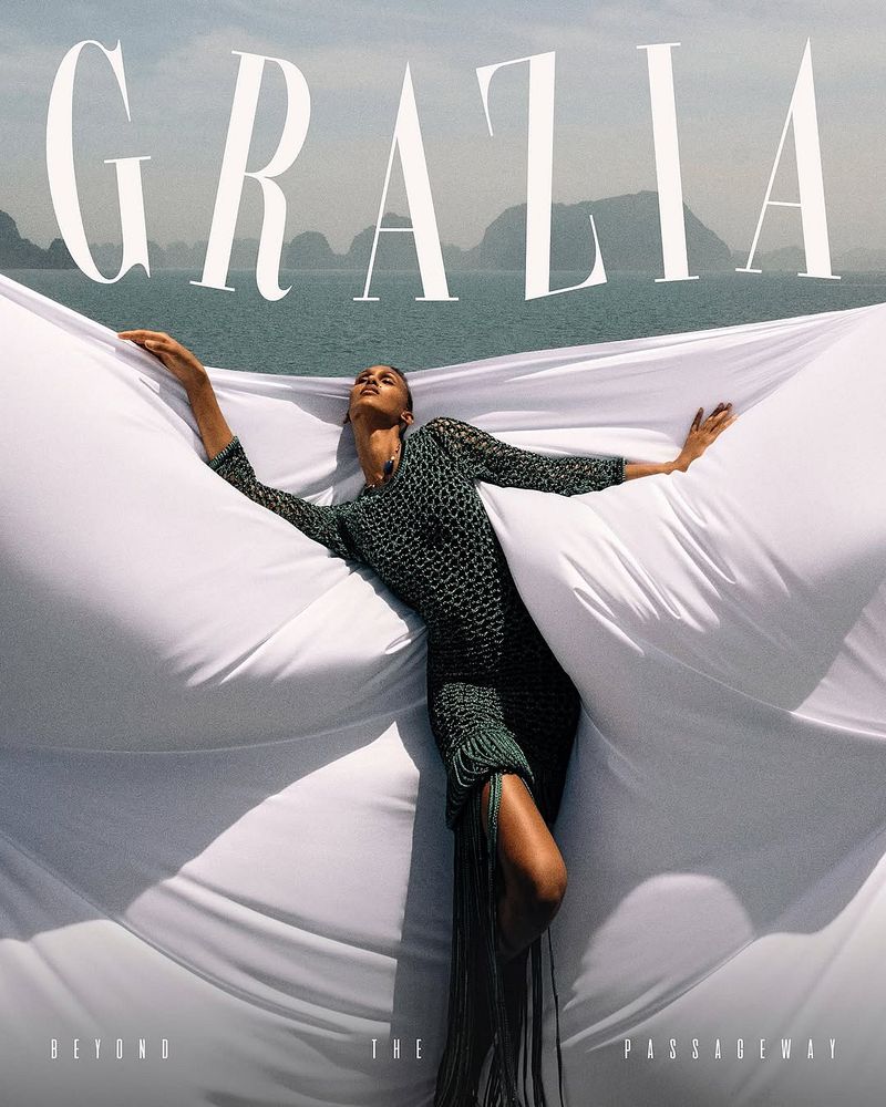 Fashion editorial featuring a woman in a green dress lying on a white fabric against a backdrop of water and islands. Magazine cover