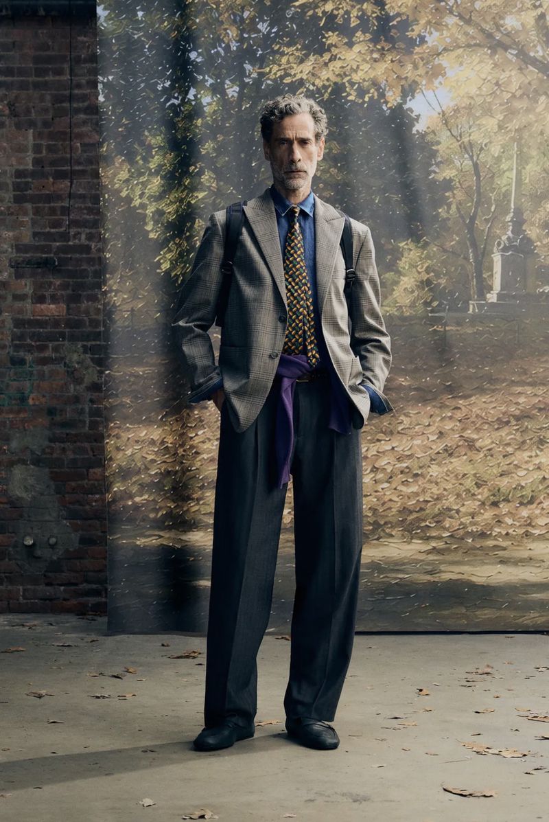 Mature man in a grey wool blazer and blue shirt with a patterned tie and purple waist wrap stands in front of a fall landscape backdrop.