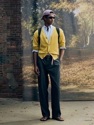 Model wearing a yellow cardigan, dark tailored trousers, purple baseball cap, sunglasses, and sandals against a scenic backdrop.
