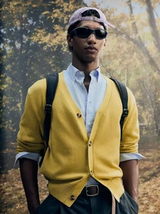 Man wearing a yellow knit cardigan over a light blue collared shirt, sunglasses, and a backwards cap in an autumn woodland setting.