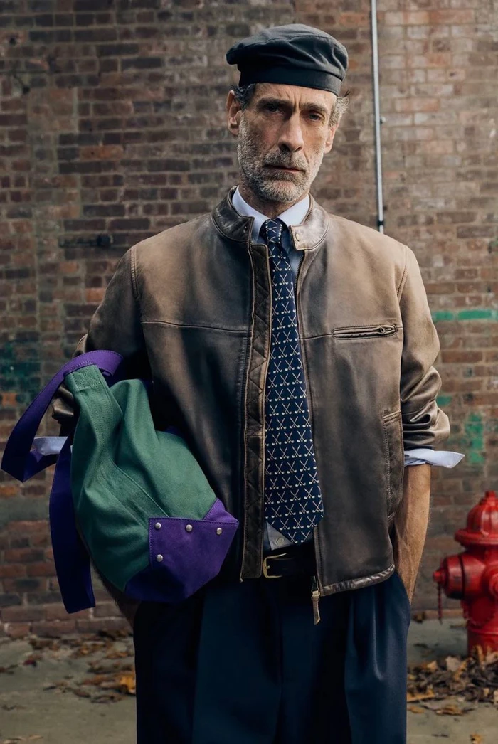 Mature man in a brown distressed leather jacket, beret, patterned tie, and dark trousers holding a green and purple tote bag.