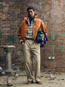 A male model wearing an orange hooded jacket, plaid shirt, and beige pleated trousers standing in front of an urban brick wall with pigeons.