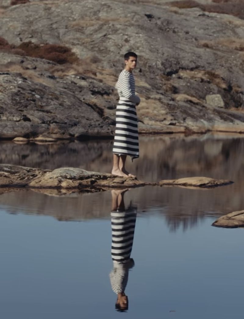 Man wearing a striped long-sleeve shirt and matching sarong-like towel standing on a rocky outcrop reflected in still water.