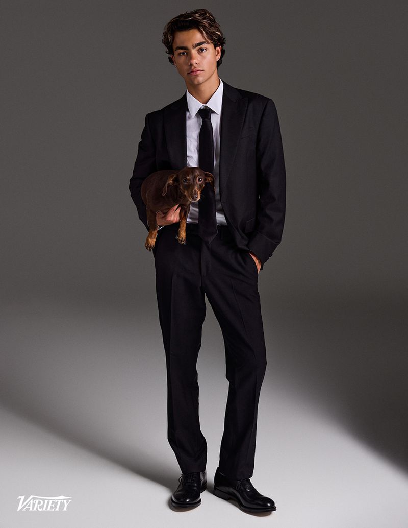 Malachi Barton wearing a black suit and tie while holding a small brown dog against a grey studio backdrop.