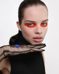 Close-up portrait of a woman with vibrant orange eye makeup and sheer black mesh gloves against a pure white studio background.