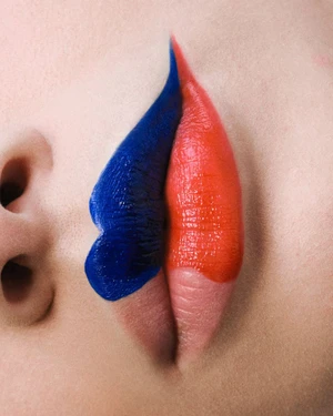 Macro view of lips painted with graphic two-tone royal blue and vibrant coral-red lipstick against pale skin.