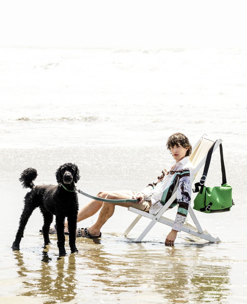 Model relaxing on a white deck chair in shallow ocean water holding the leash of a black poodle with a green shoulder bag hanging.
