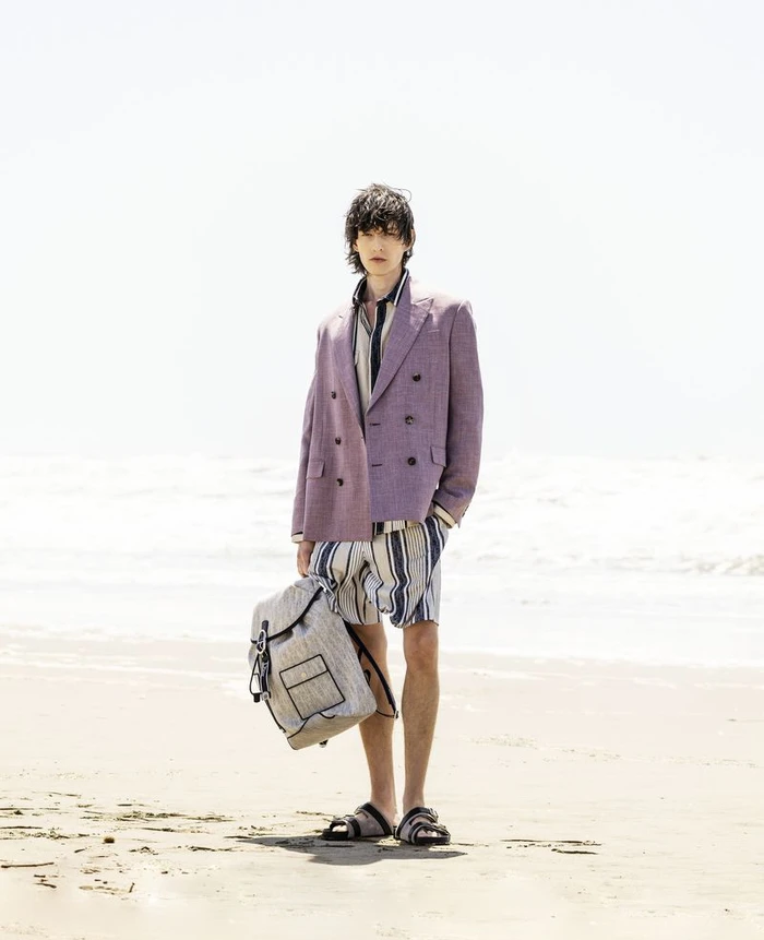 Model Erik Kettschick standing on a sandy beach wearing a purple double-breasted blazer, striped shorts, and holding a backpack.