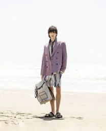 Model Erik Kettschick standing on a sandy beach wearing a purple double-breasted blazer, striped shorts, and holding a backpack.