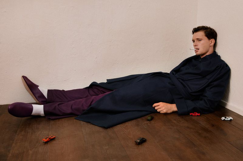Model Jakob Jokerst reclining on a wooden floor against an off-white wall in a navy robe, purple trousers, and smoking a cigarette.