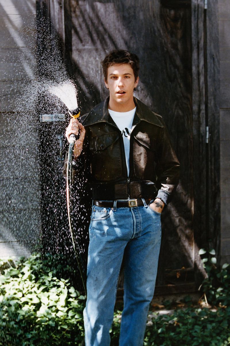A young man holding a garden hose spraying water in front of a dark wooden door while wearing a leather jacket and blue denim jeans.