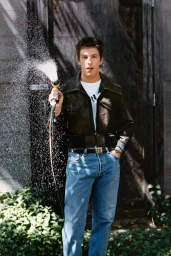 Musician Role Model holding and spraying a garden hose wearing a dark leather jacket and blue jeans in an outdoor yard.