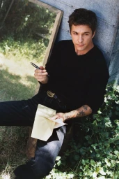 Role Model seated outdoors in a black sweater and dark jeans holding a pen and writing pad.