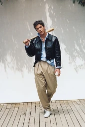 Musician Role Model posing with a wooden baseball bat in a black leather jacket and beige pleated trousers for Interview Magazine.