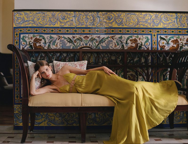 Woman in a long yellow halter dress reclining on a dark wood bench against ornate blue and yellow tiled wall.