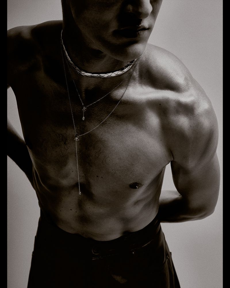 Close-up of a shirtless muscular man wearing multiple delicate silver chains around his neck against a plain grey studio background.