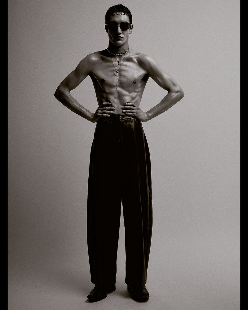 A shirtless man wearing dark sunglasses, necklace, and high-waisted wide-leg trousers stands with hands on hips against a neutral backdrop.