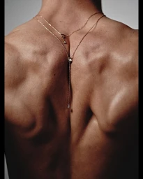 Close-up of a person's bare muscular back adorned with a delicate gold chain necklace draped down the spine.