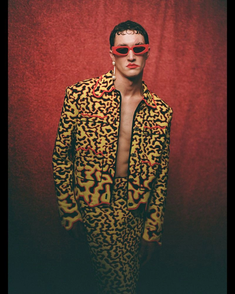 Model in a yellow patterned jacket, red angular sunglasses, and drop earring posed against a textured red studio backdrop.