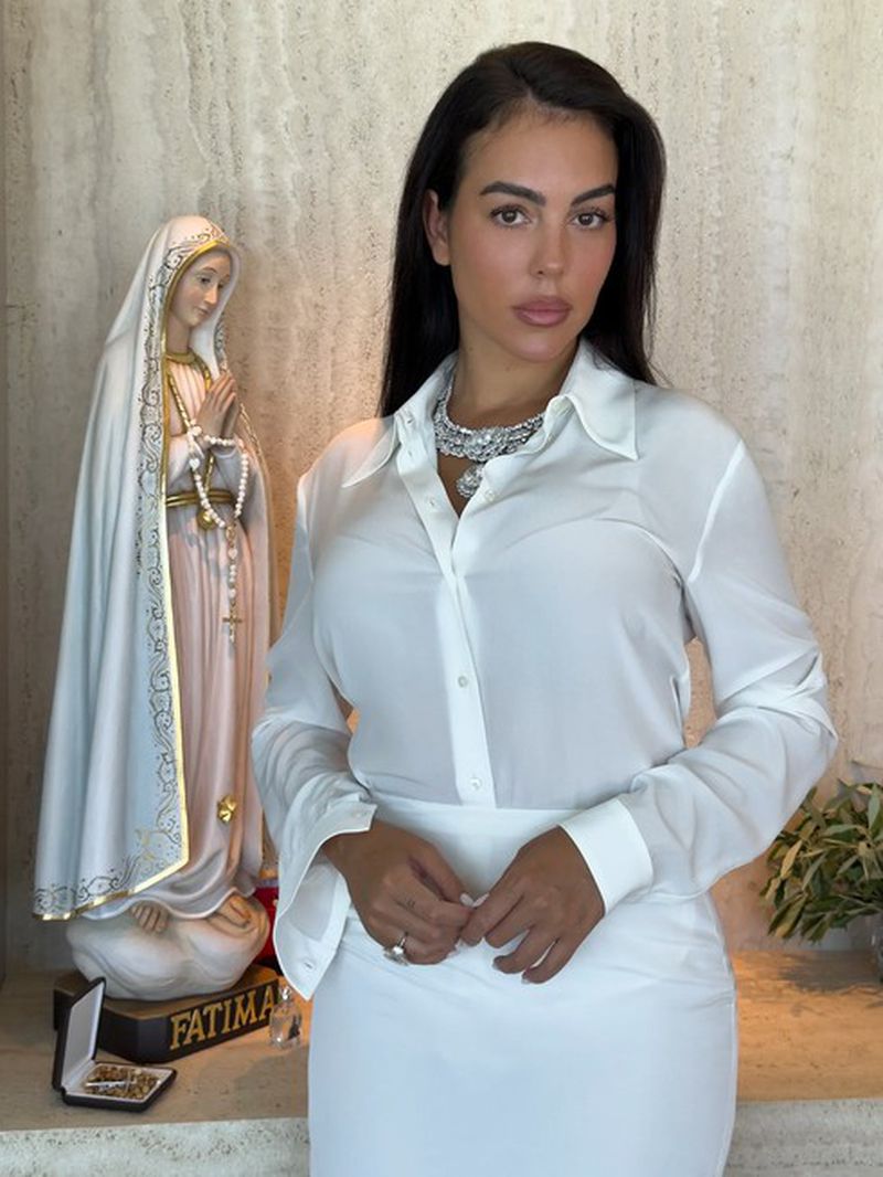 Georgina Rodriguez wearing a white button-up shirt and diamond necklace beside a statue of Our Lady of Fatima.