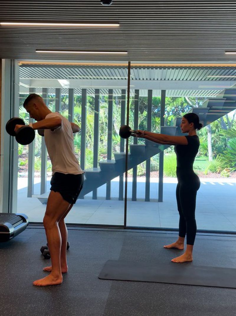 Cristiano Ronaldo and Georgina Rodriguez exercising in a bright modern home gym with floor to ceiling glass walls and a staircase.