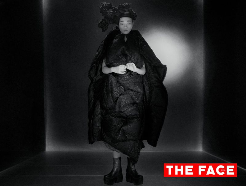 Model wearing a voluminous sculptural black coat, elaborate floral headpiece, and platform boots in a dark studio setting.